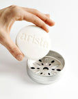 Hand holding a white 'arista' strengthening shampoo bar over a silver shampoo holder box on a light background