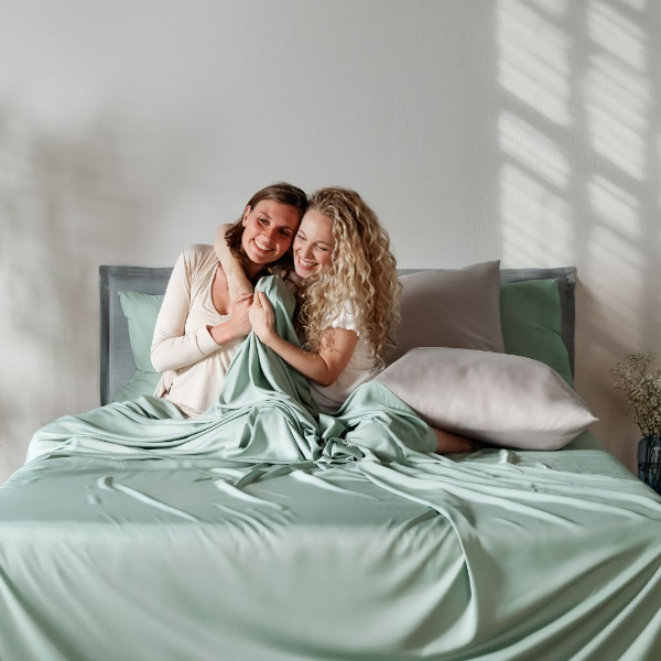 Two women hugging under a light green duvet on a bed with white walls and a window in the background.