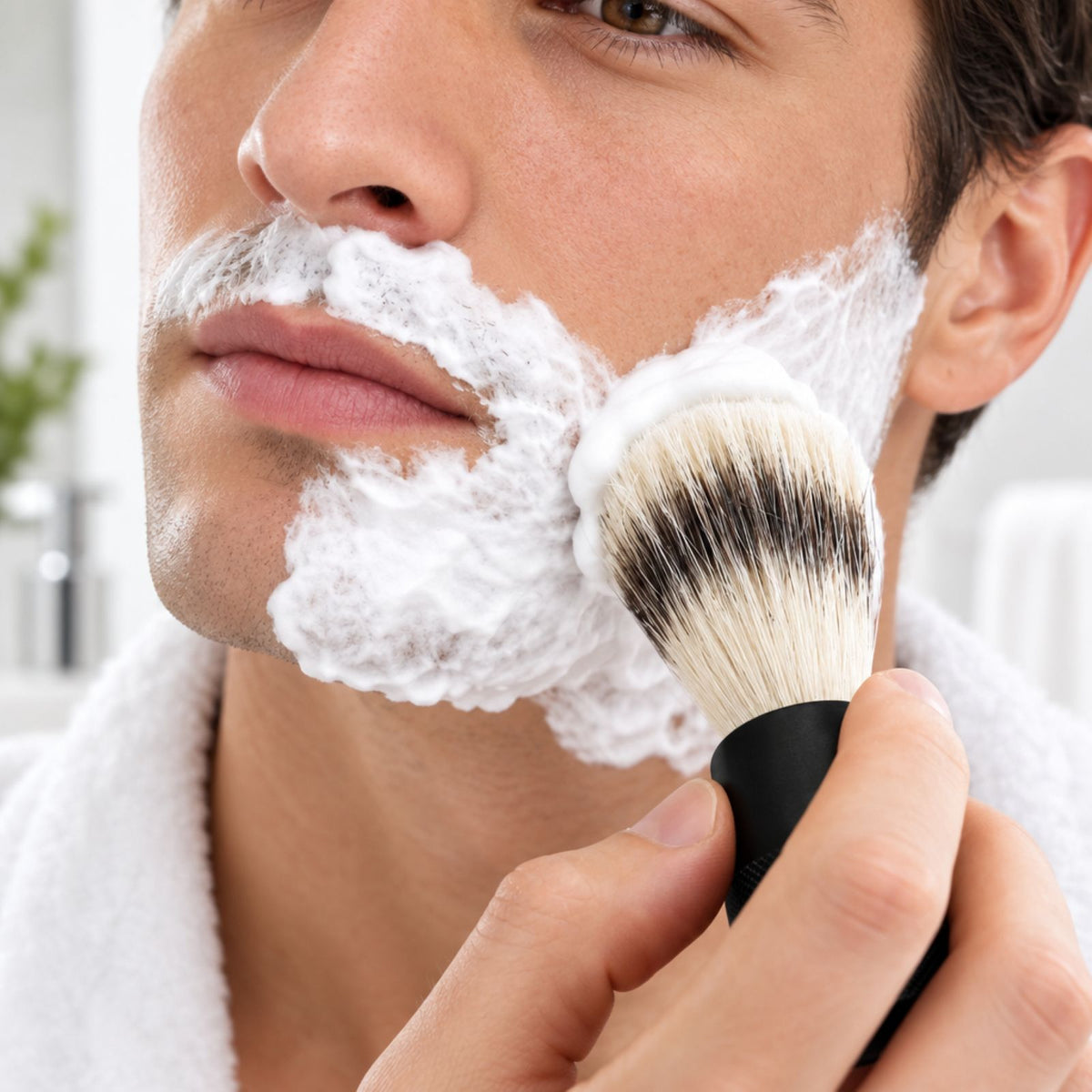 A close-up of a man shaving his face with white shaving foam, holding a bambaw metal shaving brush with black and white bristles and a black handle.