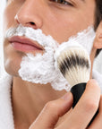 A close-up of a man shaving his face with white shaving foam, holding a bambaw metal shaving brush with black and white bristles and a black handle.