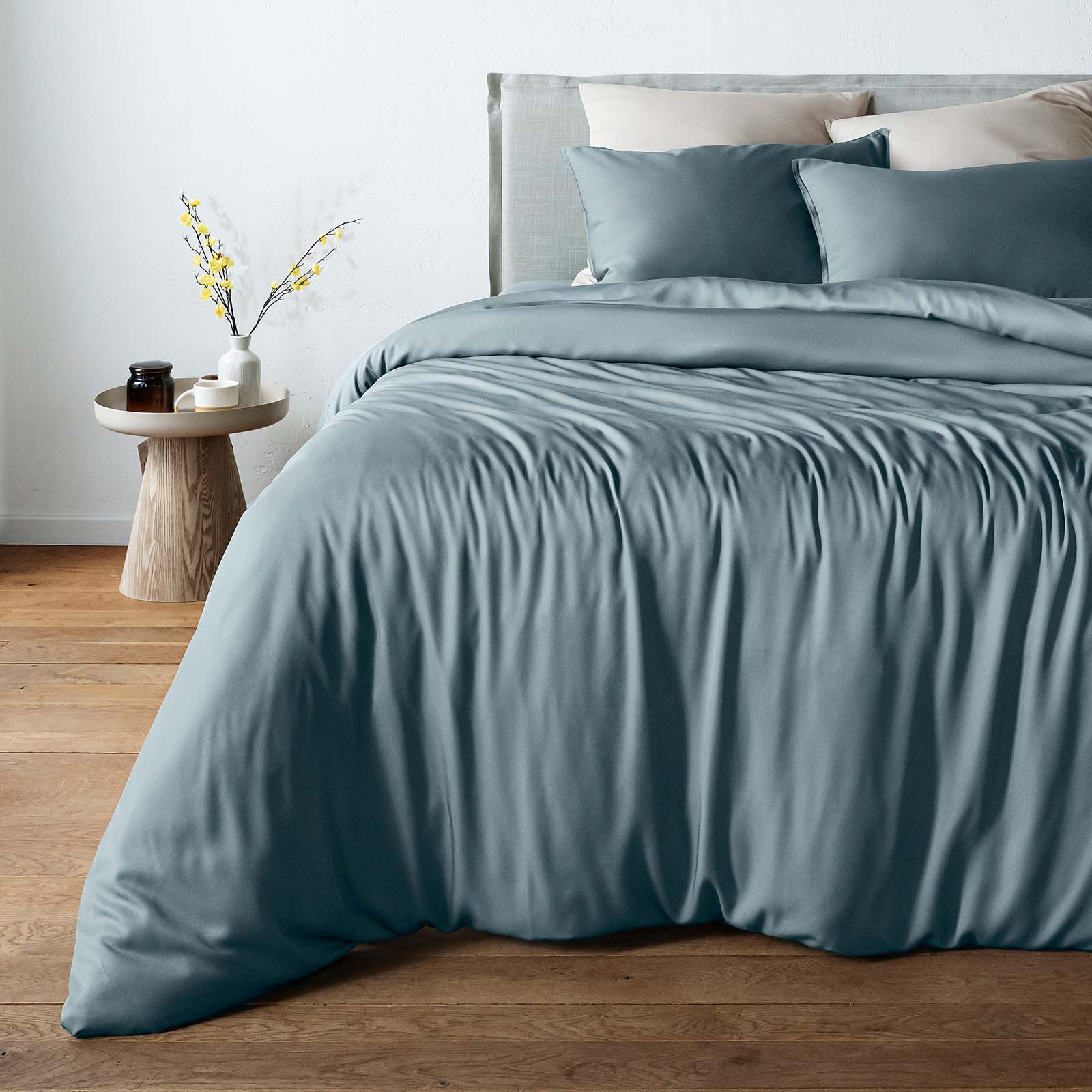 A bed dressed in a dusty blue bamboo duvet cover and matching pillow shams, styled in a bedroom with a wooden floor, a light wood side table, and neutral wall tones.